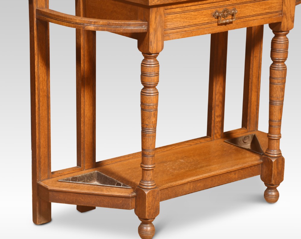 An antique carved oak hall stand featuring a central circular mirror, upper coat hooks, a small shelf above a single drawer, and side umbrella stands with lower trays. View V