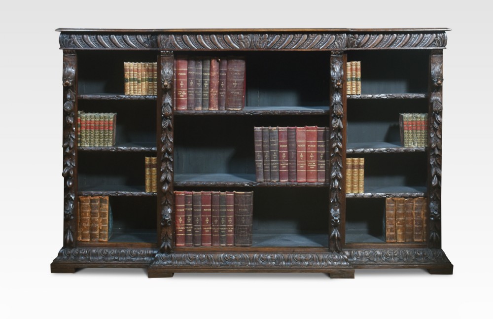 A low, dark carved oak open bookcase with a breakfront design, featuring three shelf sections separated by carved pilasters decorated with foliate and mask motifs. View II