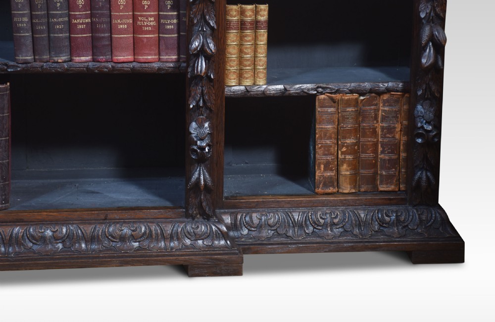 A low, dark carved oak open bookcase with a breakfront design, featuring three shelf sections separated by carved pilasters decorated with foliate and mask motifs. View V