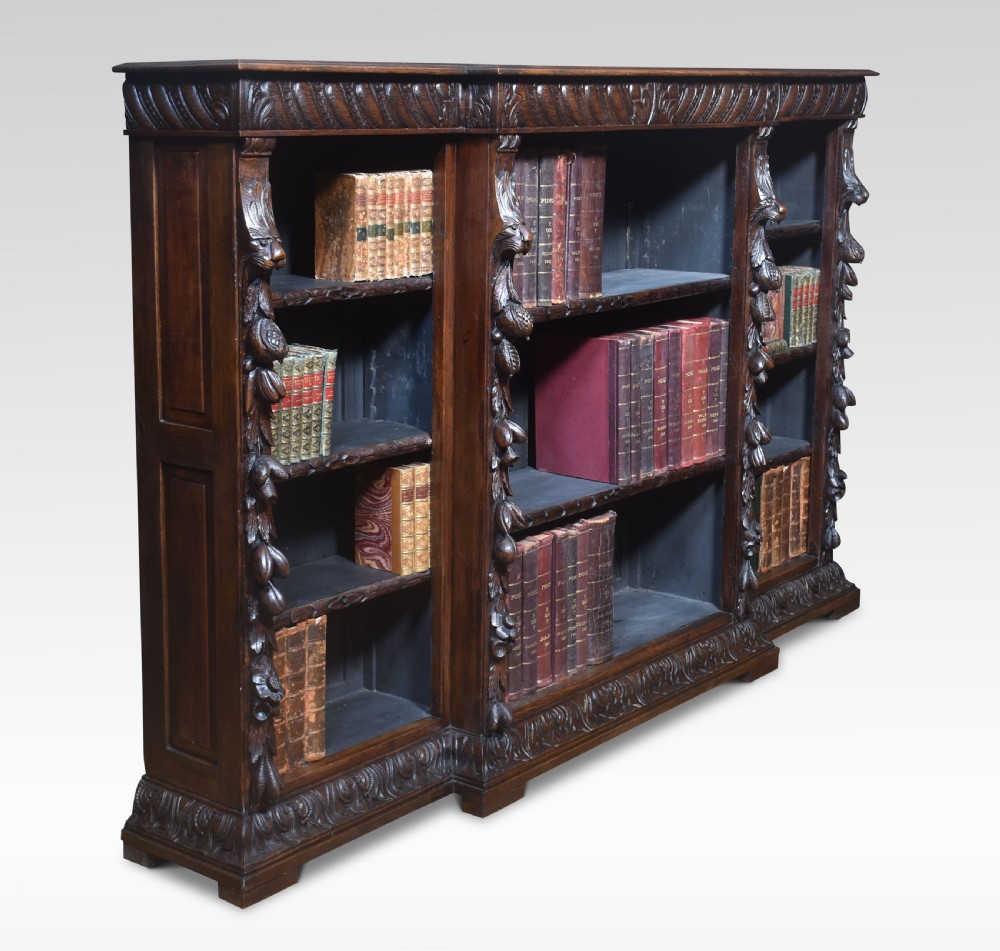 A low, dark carved oak open bookcase with a breakfront design, featuring three shelf sections separated by carved pilasters decorated with foliate and mask motifs. View VI