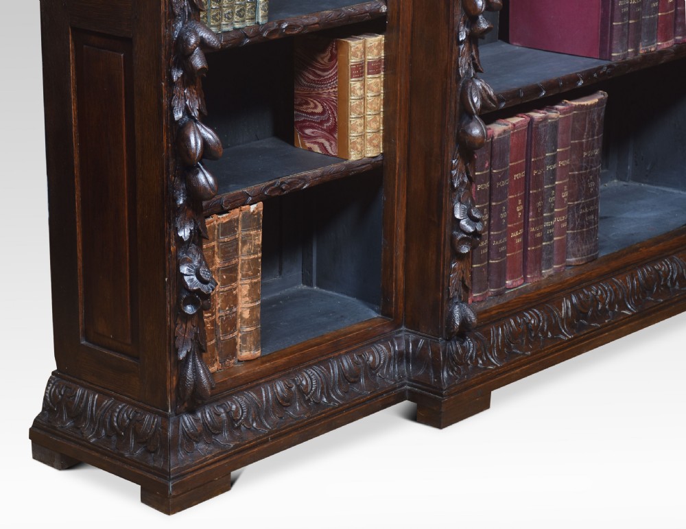 A low, dark carved oak open bookcase with a breakfront design, featuring three shelf sections separated by carved pilasters decorated with foliate and mask motifs. View IX