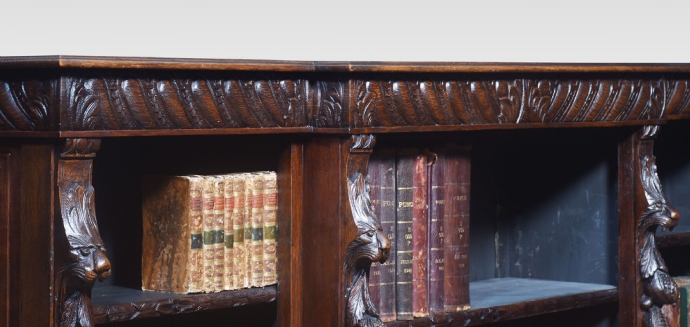A low, dark carved oak open bookcase with a breakfront design, featuring three shelf sections separated by carved pilasters decorated with foliate and mask motifs. View X