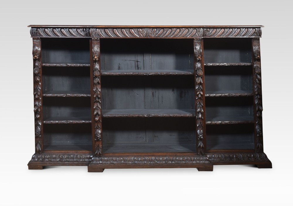 A low, dark carved oak open bookcase with a breakfront design, featuring three shelf sections separated by carved pilasters decorated with foliate and mask motifs. View XI