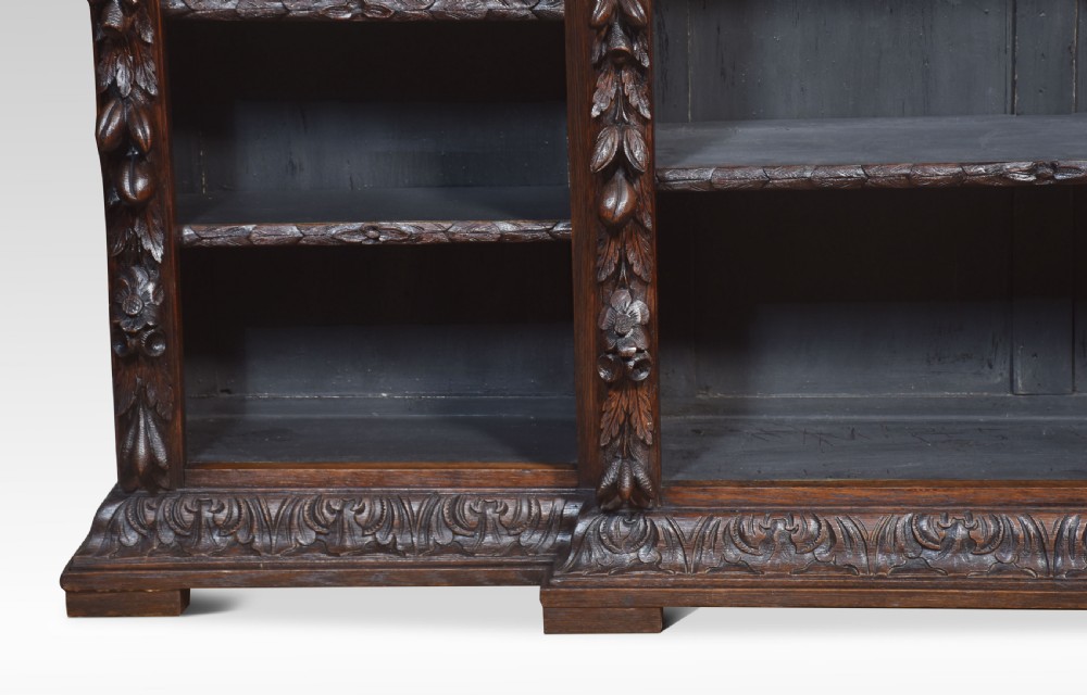 A low, dark carved oak open bookcase with a breakfront design, featuring three shelf sections separated by carved pilasters decorated with foliate and mask motifs. View XII