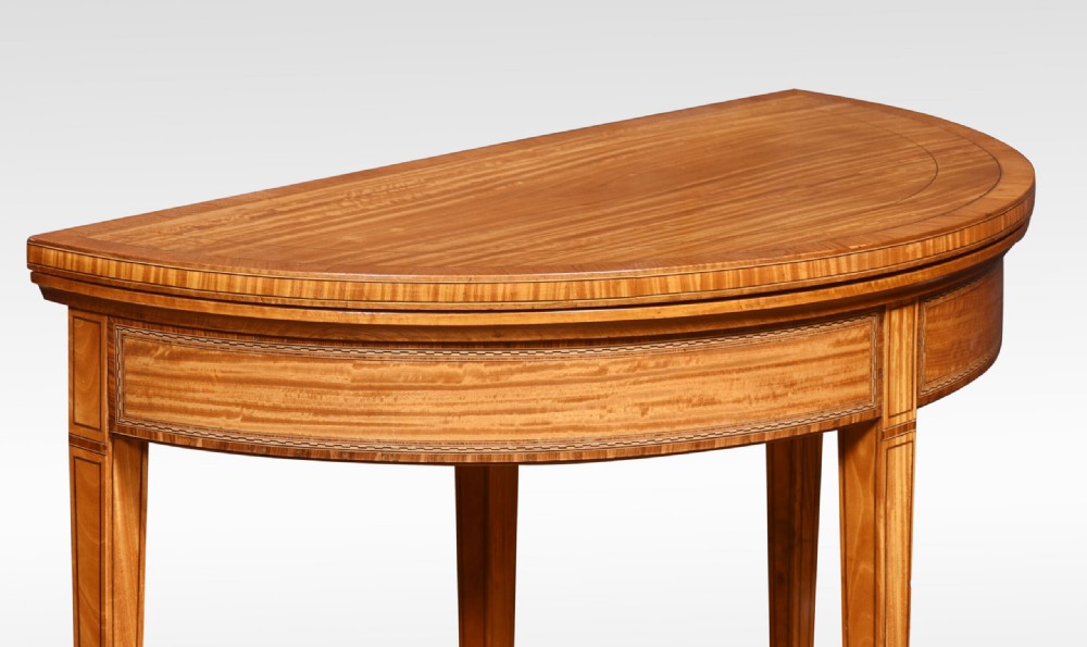 An antique semi-circular satinwood card table featuring inlaid crossbanding, tapered square legs, and small brass castors. View IV