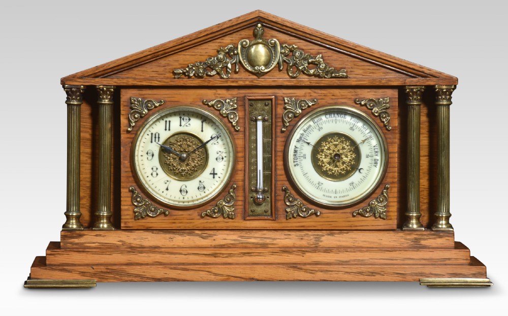 An architectural oak desk clock featuring a triangular pediment top, brass fluted columns, a central thermometer, and twin dials for a clock and barometer.
