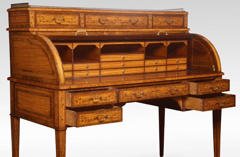 A Sheraton Revival satinwood cylinder bureau featuring detailed floral marquetry inlays, a curved roll-top front, brass gallery, and multiple drawers raised on tapered square legs. View VI