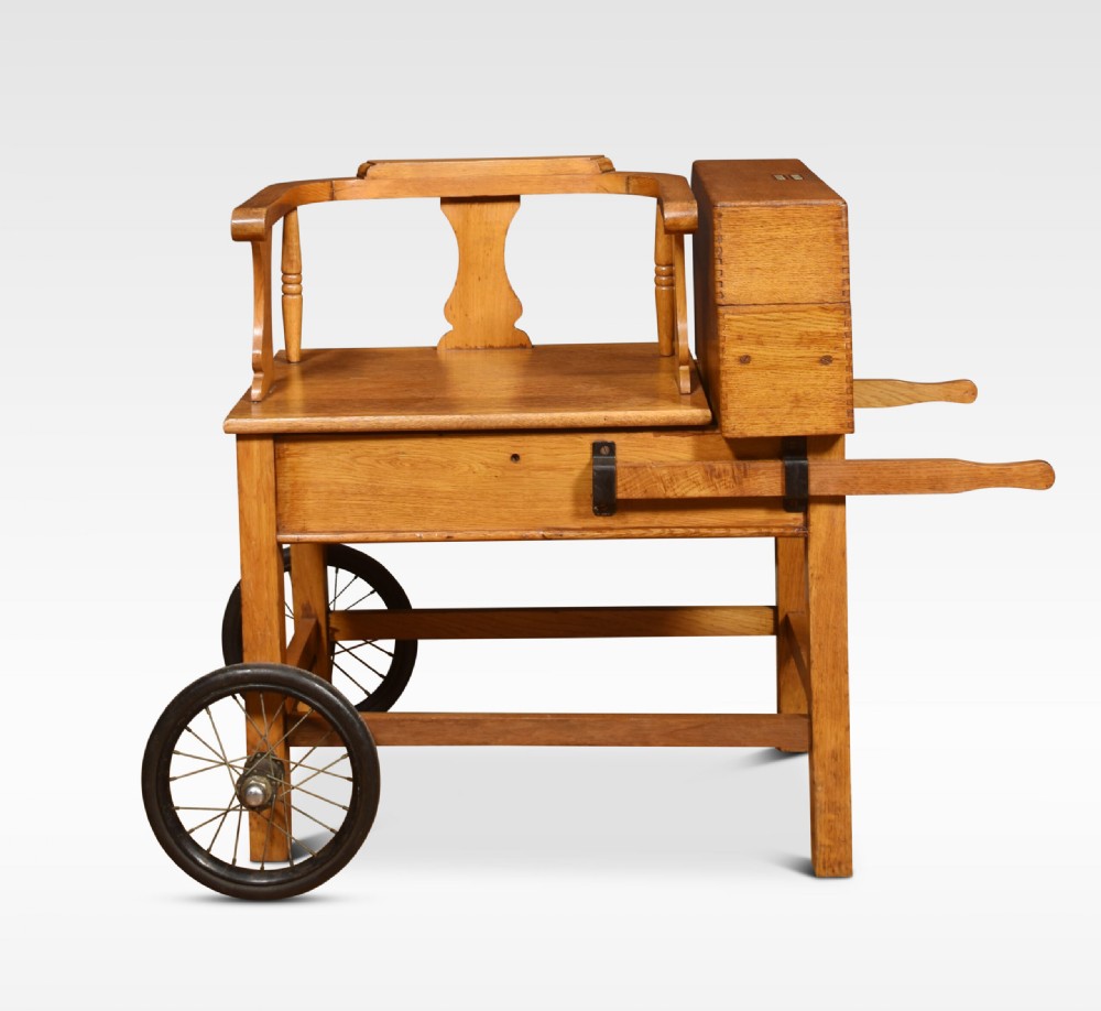 An antique oak doctors weighing scale chair with two spoked wheels, a wooden armchair frame, and a side-mounted brass weighing beam mechanism. View II