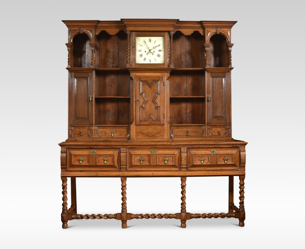 A Jacobean style oak dresser featuring geometric panel moulding, a central integrated clock, open upper shelves, and six barley twist legs.