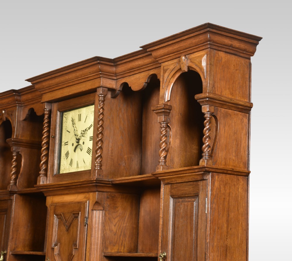 A Jacobean style oak dresser featuring geometric panel moulding, a central integrated clock, open upper shelves, and six barley twist legs. View VII