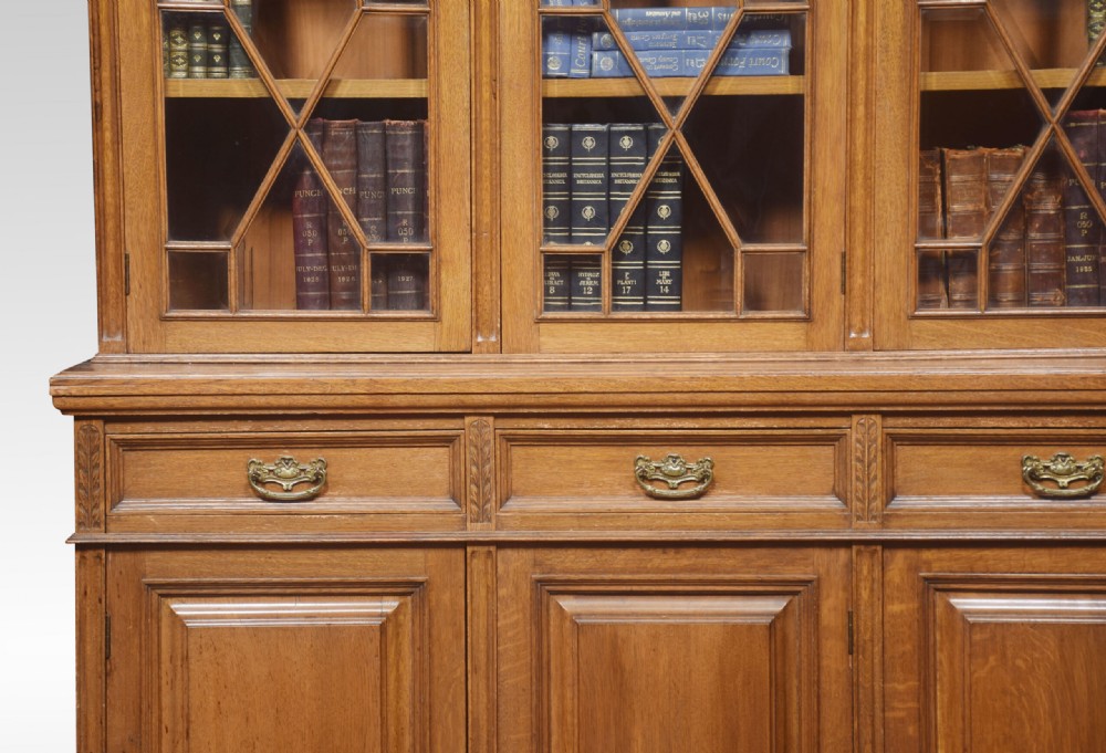A large warm brown oak bookcase featuring three upper glazed doors with geometric astragal glazing, three drawers, and three panelled cupboard doors below. View VI