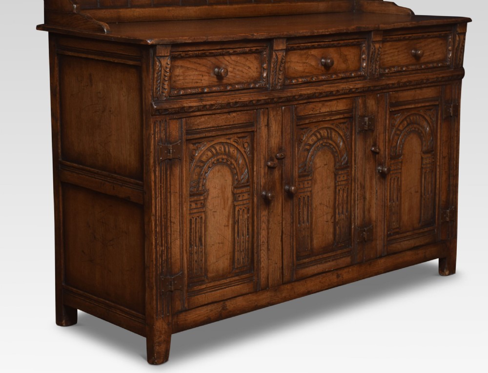 A Jacobean style dark oak dresser featuring a tiered open plate rack above three drawers and three carved panelled cupboard doors. View V