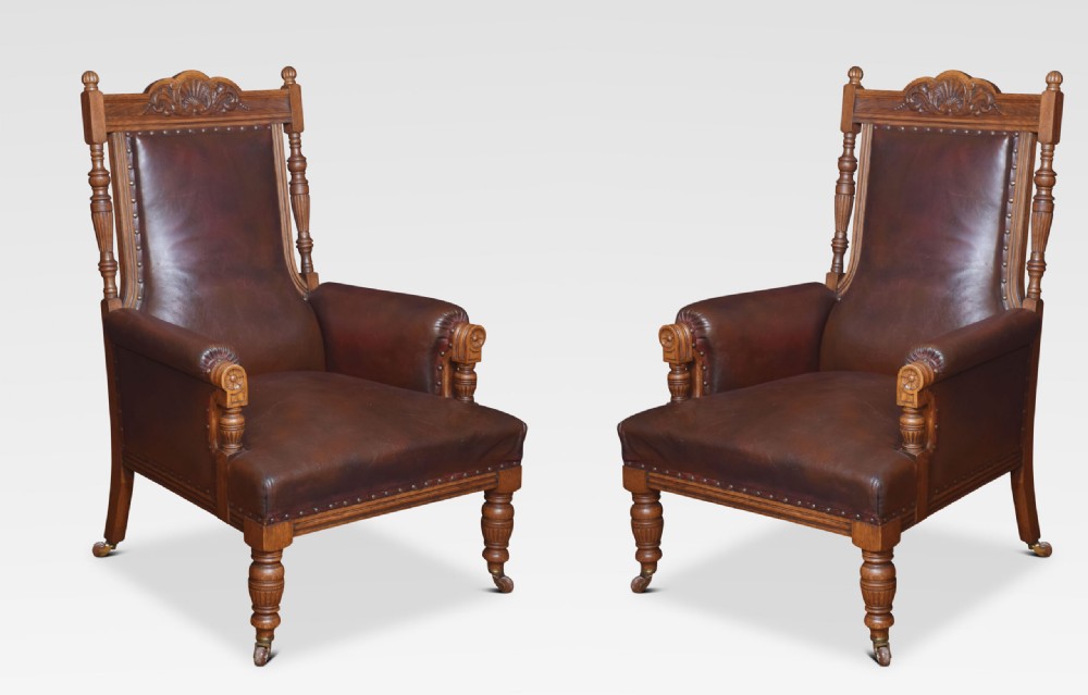 A pair of antique Victorian carved oak library armchairs upholstered in dark brown leather with brass studding and turned front legs on castors.