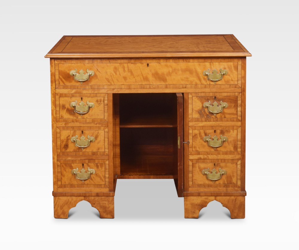 A George III style satinwood kneehole desk featuring crossbanded drawers, brass batwing handles, a central recessed cupboard, and bracket feet.
