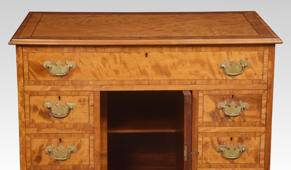 A George III style satinwood kneehole desk featuring crossbanded drawers, brass batwing handles, a central recessed cupboard, and bracket feet. View III