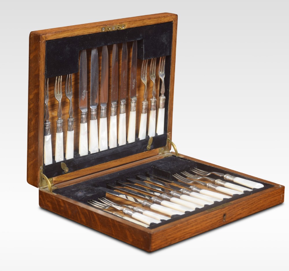 An open wooden oak case displaying a structured set of silver-plated knives and forks with mother-of-pearl handles set against dark velvet lining.