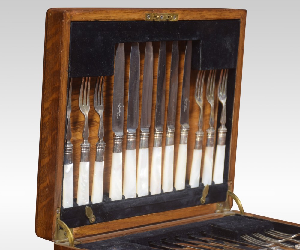 An open wooden oak case displaying a structured set of silver-plated knives and forks with mother-of-pearl handles set against dark velvet lining. View II