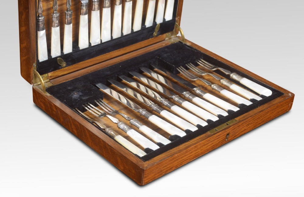 An open wooden oak case displaying a structured set of silver-plated knives and forks with mother-of-pearl handles set against dark velvet lining. View IV