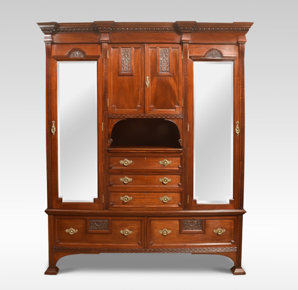 An antique carved mahogany compactum wardrobe featuring twin full-length mirrored doors flanking a central cupboard section, open niche, and multiple drawers with brass hardware.