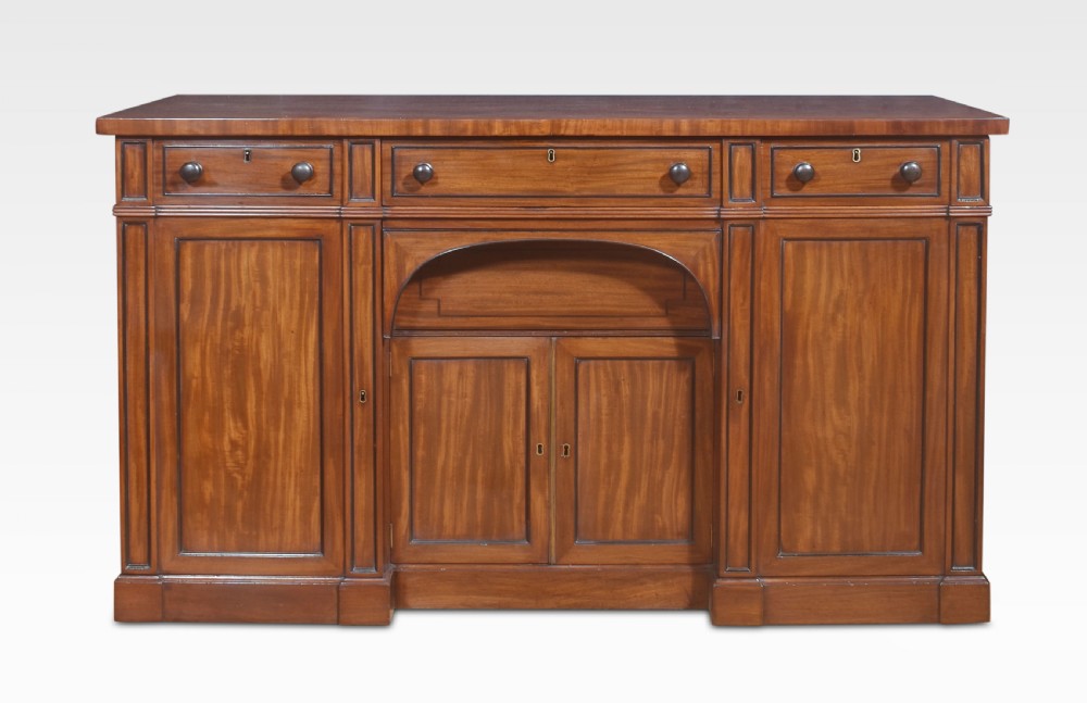 A Regency period warm brown mahogany sideboard featuring four cupboard doors, three top drawers, turned wooden knobs, and an arched central alcove.