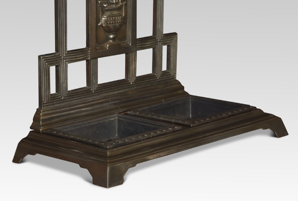 A dark cast iron umbrella stand featuring a triangular pediment top, central floral urn relief panel, upper retaining bar, and a double-compartment drip tray at the base. View II