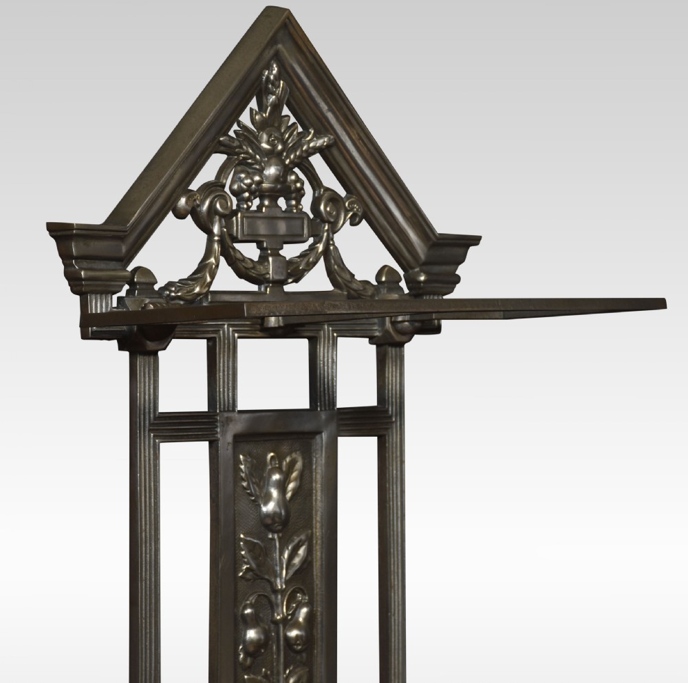 A dark cast iron umbrella stand featuring a triangular pediment top, central floral urn relief panel, upper retaining bar, and a double-compartment drip tray at the base. View IV