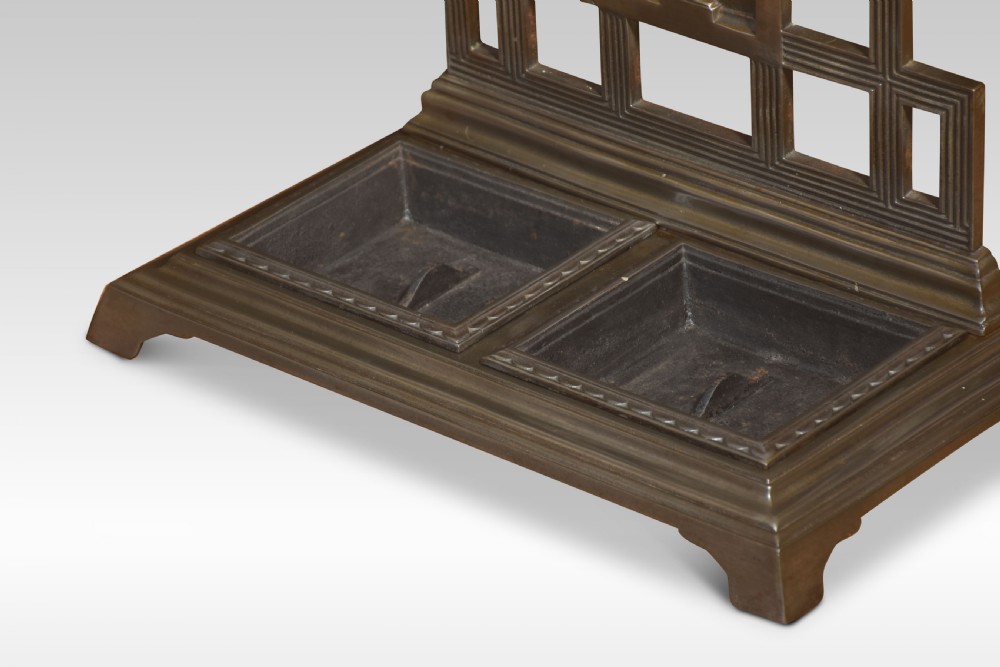 A dark cast iron umbrella stand featuring a triangular pediment top, central floral urn relief panel, upper retaining bar, and a double-compartment drip tray at the base. View VIII