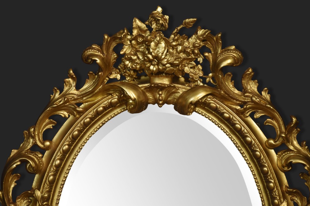 An oval wall mirror featuring a carved gold gilded frame decorated with scrolling acanthus leaves and a central floral crest. View II
