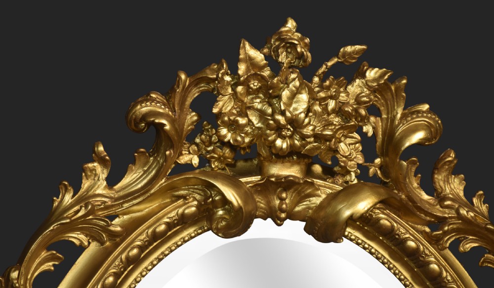 An oval wall mirror featuring a carved gold gilded frame decorated with scrolling acanthus leaves and a central floral crest. View V