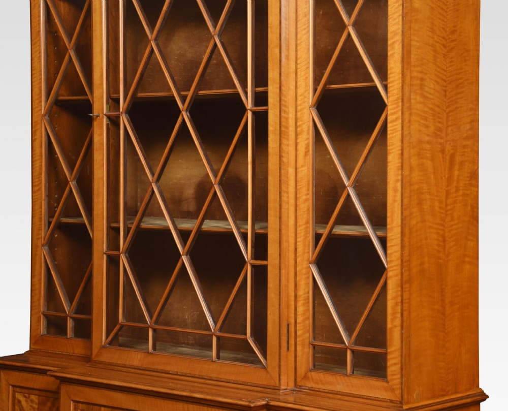 A tall Sheraton Revival satinwood breakfront bookcase featuring geometric astragal glazed upper doors above panelled lower cupboards. View V