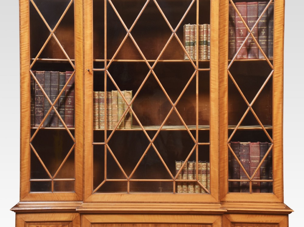 A tall Sheraton Revival satinwood breakfront bookcase featuring geometric astragal glazed upper doors above panelled lower cupboards. View VII