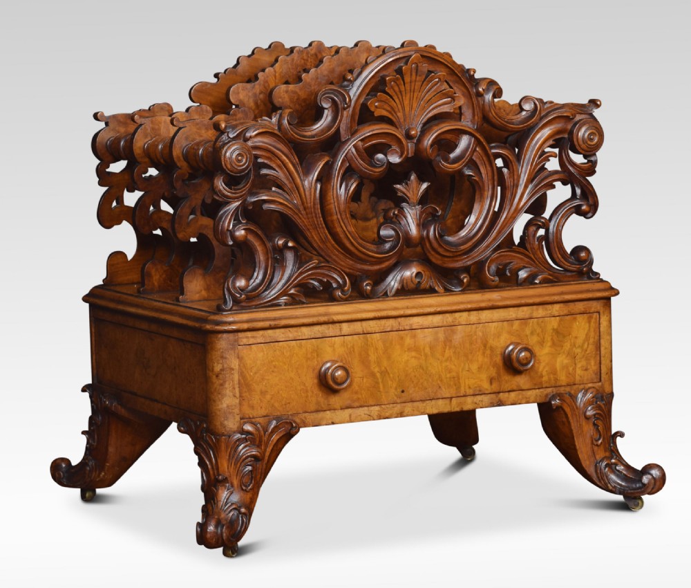 A carved Victorian figured walnut Canterbury featuring three carved scrollwork dividers, a single base drawer with wooden knobs, and cabriole legs on brass castors.