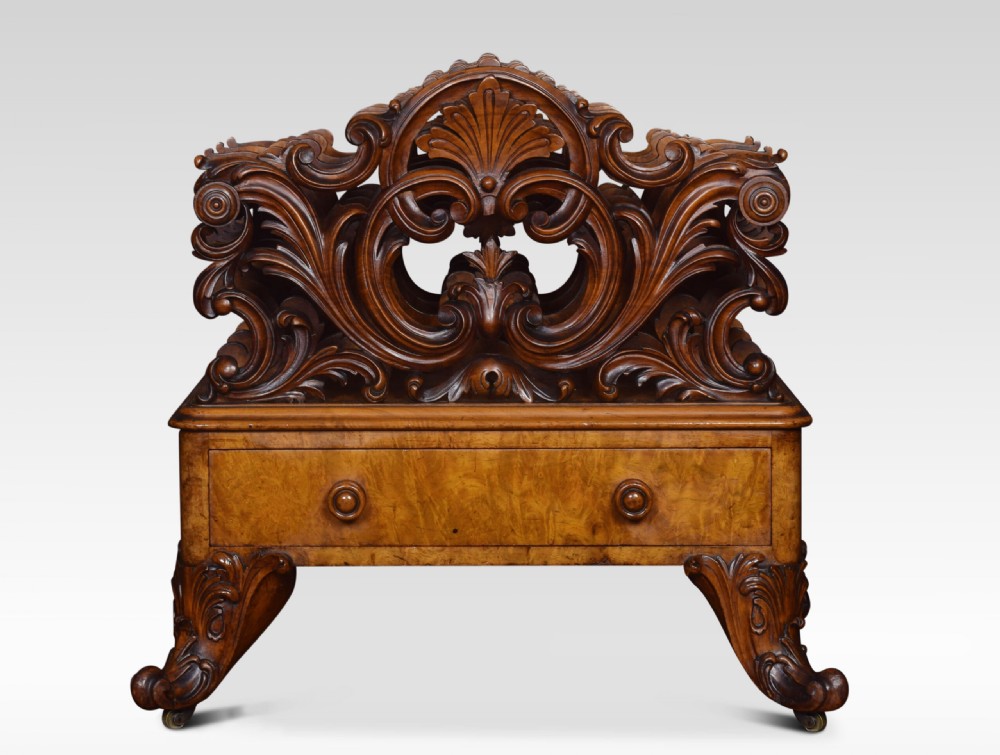 A carved Victorian figured walnut Canterbury featuring three carved scrollwork dividers, a single base drawer with wooden knobs, and cabriole legs on brass castors. View II