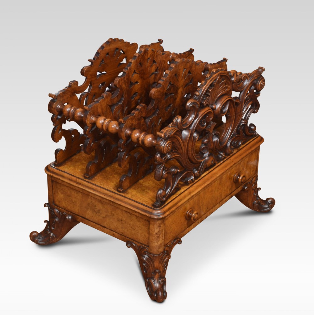 A carved Victorian figured walnut Canterbury featuring three carved scrollwork dividers, a single base drawer with wooden knobs, and cabriole legs on brass castors. View IV