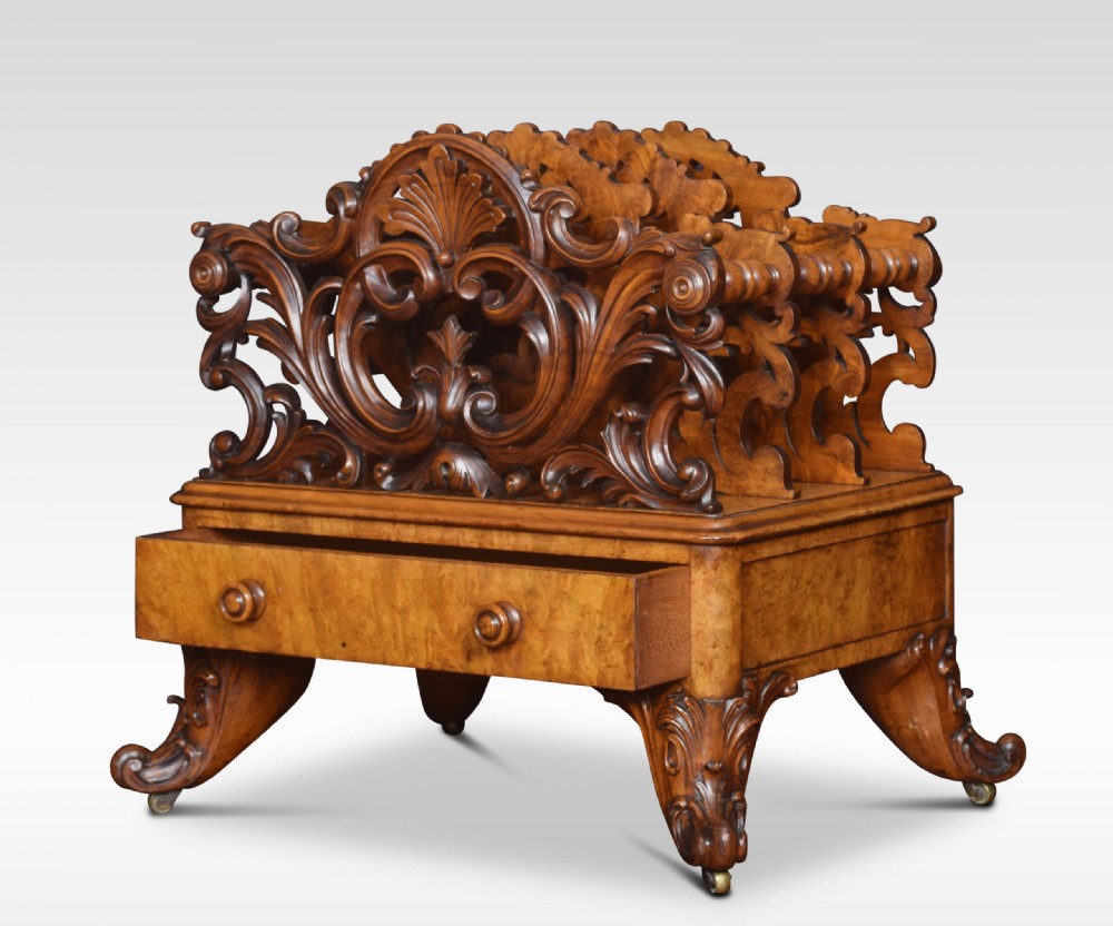 A carved Victorian figured walnut Canterbury featuring three carved scrollwork dividers, a single base drawer with wooden knobs, and cabriole legs on brass castors. View VI