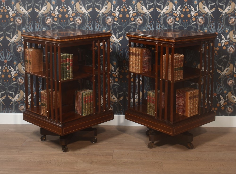A pair of Edwardian inlaid mahogany revolving bookcases featuring square tops with satinwood crossbanding, vertical slat sides, and rotating bases on castors.