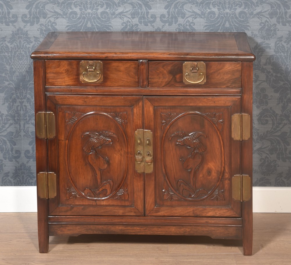 A warm brown carved Chinese wooden cupboard featuring two top drawers, brass hardware, and panelled doors depicting bird motifs.