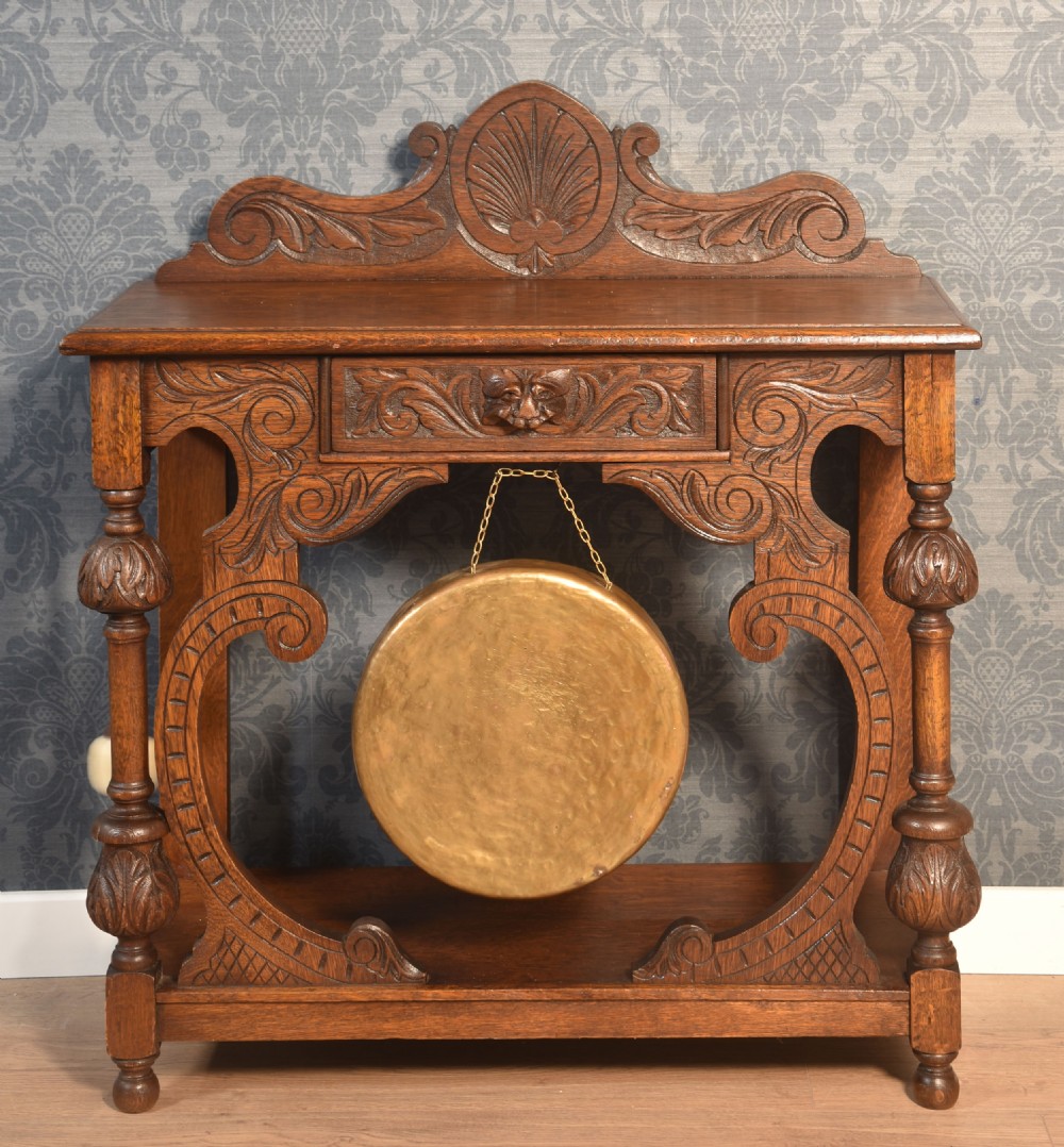 A carved dark oak hall table with an integrated suspended brass dinner gong, scrolled supports, turned legs, and an ornamental carved cresting.