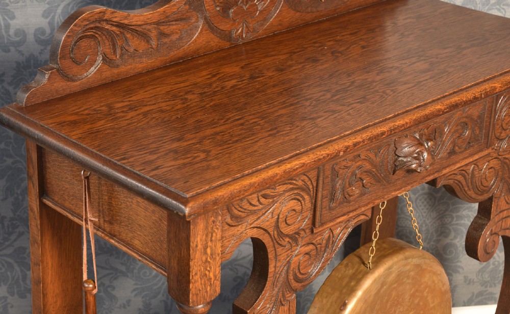 A carved dark oak hall table with an integrated suspended brass dinner gong, scrolled supports, turned legs, and an ornamental carved cresting. View VI