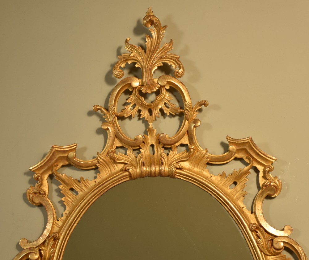 An ornate oval wall mirror featuring an intricately carved giltwood frame decorated with scrolling acanthus leaves and a pierced crest. View VI