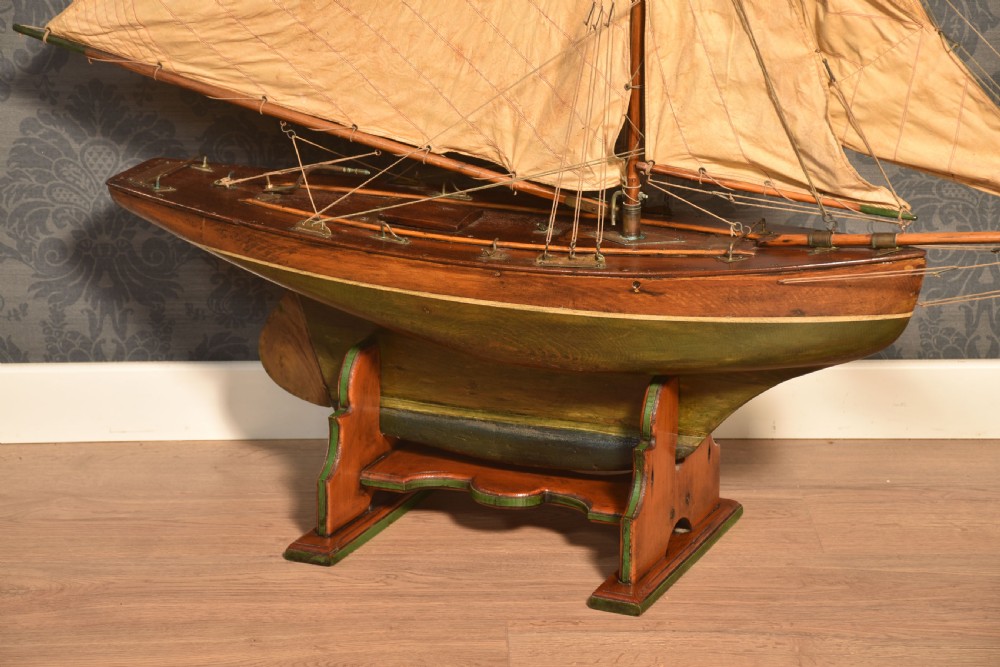 A 19th-century wooden pond yacht featuring cream canvas sails, a carved brown hull with green painted keel accents, and a wooden display stand. View IV