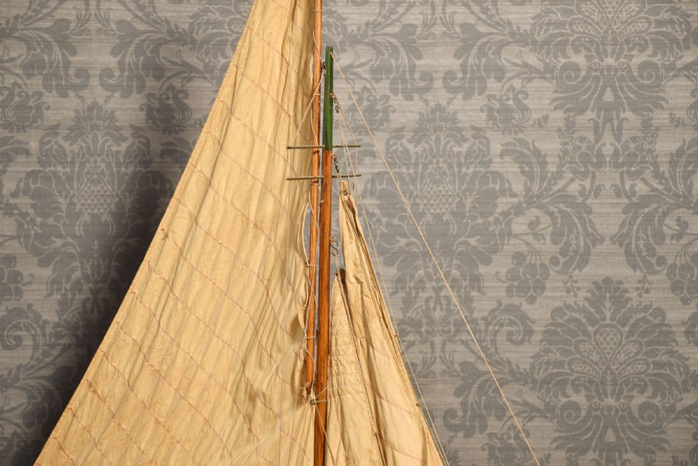 A 19th-century wooden pond yacht featuring cream canvas sails, a carved brown hull with green painted keel accents, and a wooden display stand. View VI