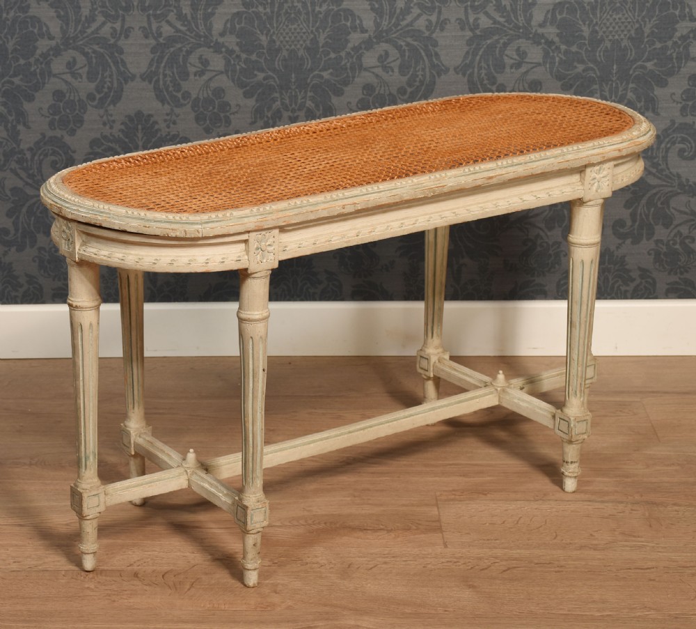 An oval window seat featuring a woven cane top set within a painted off-white wooden frame with fluted turned legs and carved rosettes.