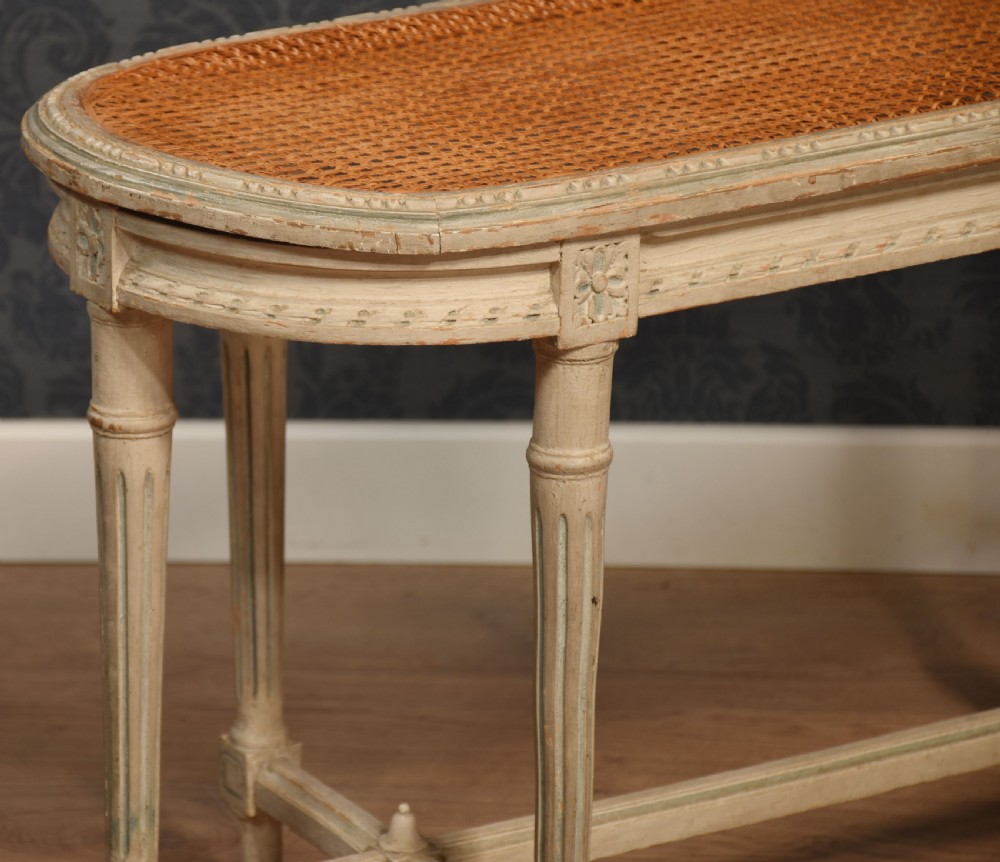 An oval window seat featuring a woven cane top set within a painted off-white wooden frame with fluted turned legs and carved rosettes. View III