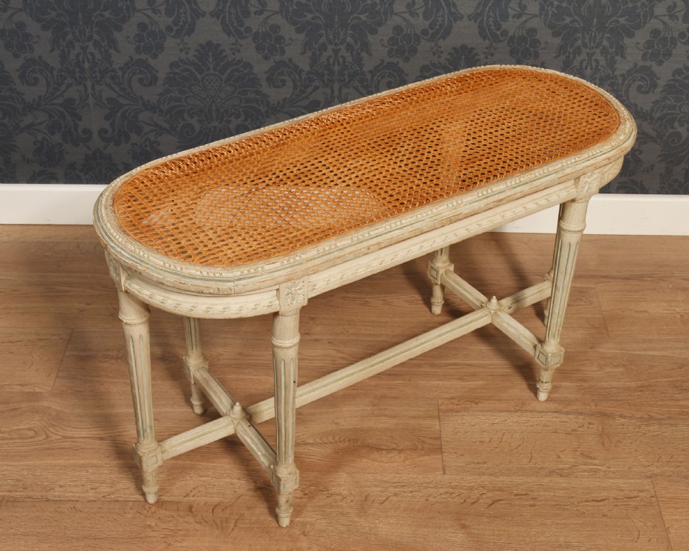 An oval window seat featuring a woven cane top set within a painted off-white wooden frame with fluted turned legs and carved rosettes. View V