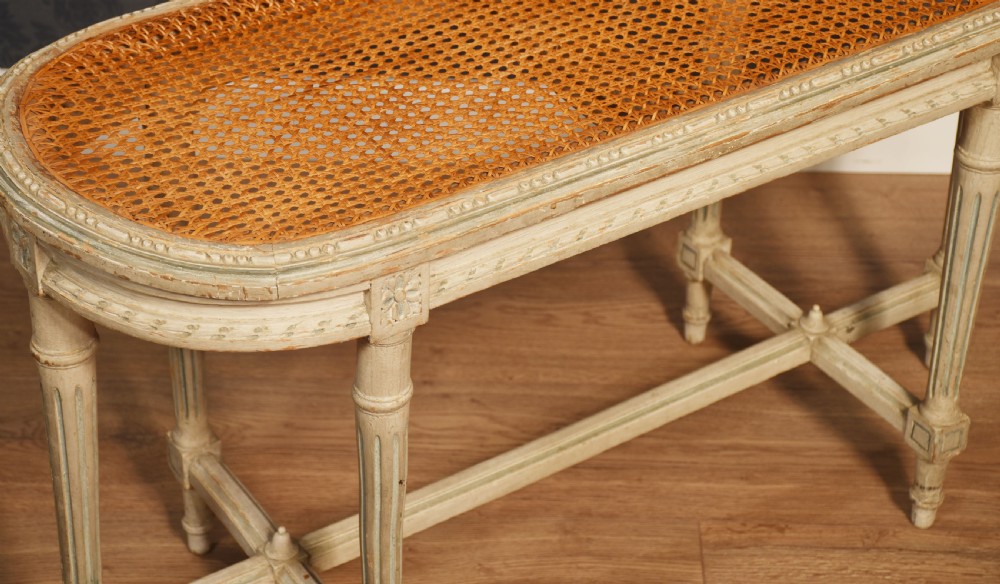 An oval window seat featuring a woven cane top set within a painted off-white wooden frame with fluted turned legs and carved rosettes. View VII