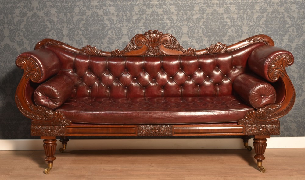 An early 19th-century deep-buttoned burgundy leather scroll-end settee with a carved mahogany frame, bolster cushions, and turned legs on brass castors.