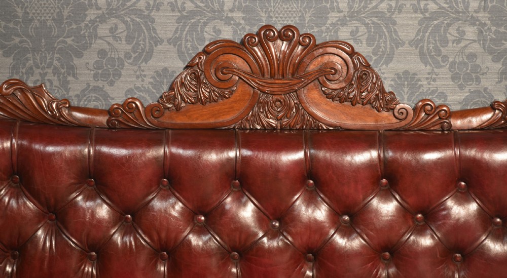 An early 19th-century deep-buttoned burgundy leather scroll-end settee with a carved mahogany frame, bolster cushions, and turned legs on brass castors. View II