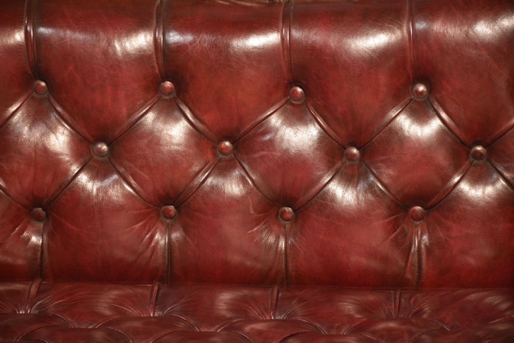An early 19th-century deep-buttoned burgundy leather scroll-end settee with a carved mahogany frame, bolster cushions, and turned legs on brass castors. View V