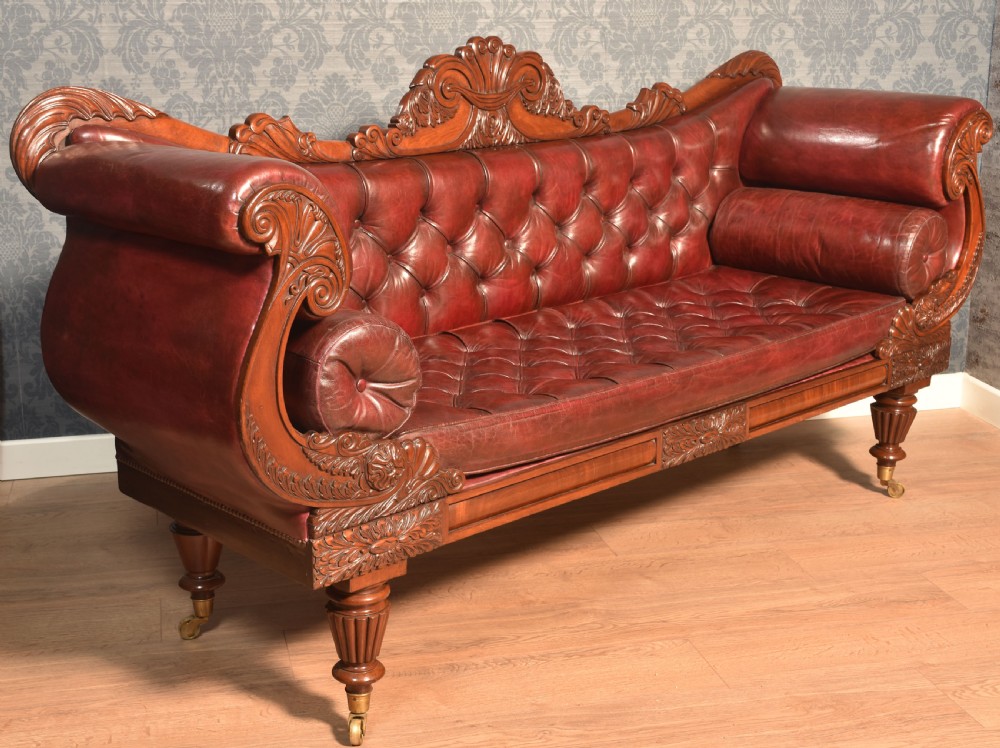 An early 19th-century deep-buttoned burgundy leather scroll-end settee with a carved mahogany frame, bolster cushions, and turned legs on brass castors. View VI
